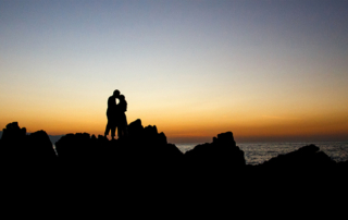 Couple kissing on a rocky shore at sunset.